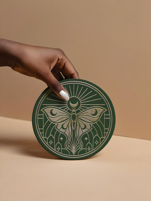 A round, dark hunter green ceramic drink coaster with a non-slip cork back. The top surface features an intricate, engraved-style celestial moth design in a light beige or off-white color. The luna moth is centered under a crescent moon and a radiant sunburst. The design is symmetrical and includes stars and geometric line art, evoking a gothic, witchy, and spiritual aesthetic. The coaster has a matte finish and is designed as a piece of functional art.